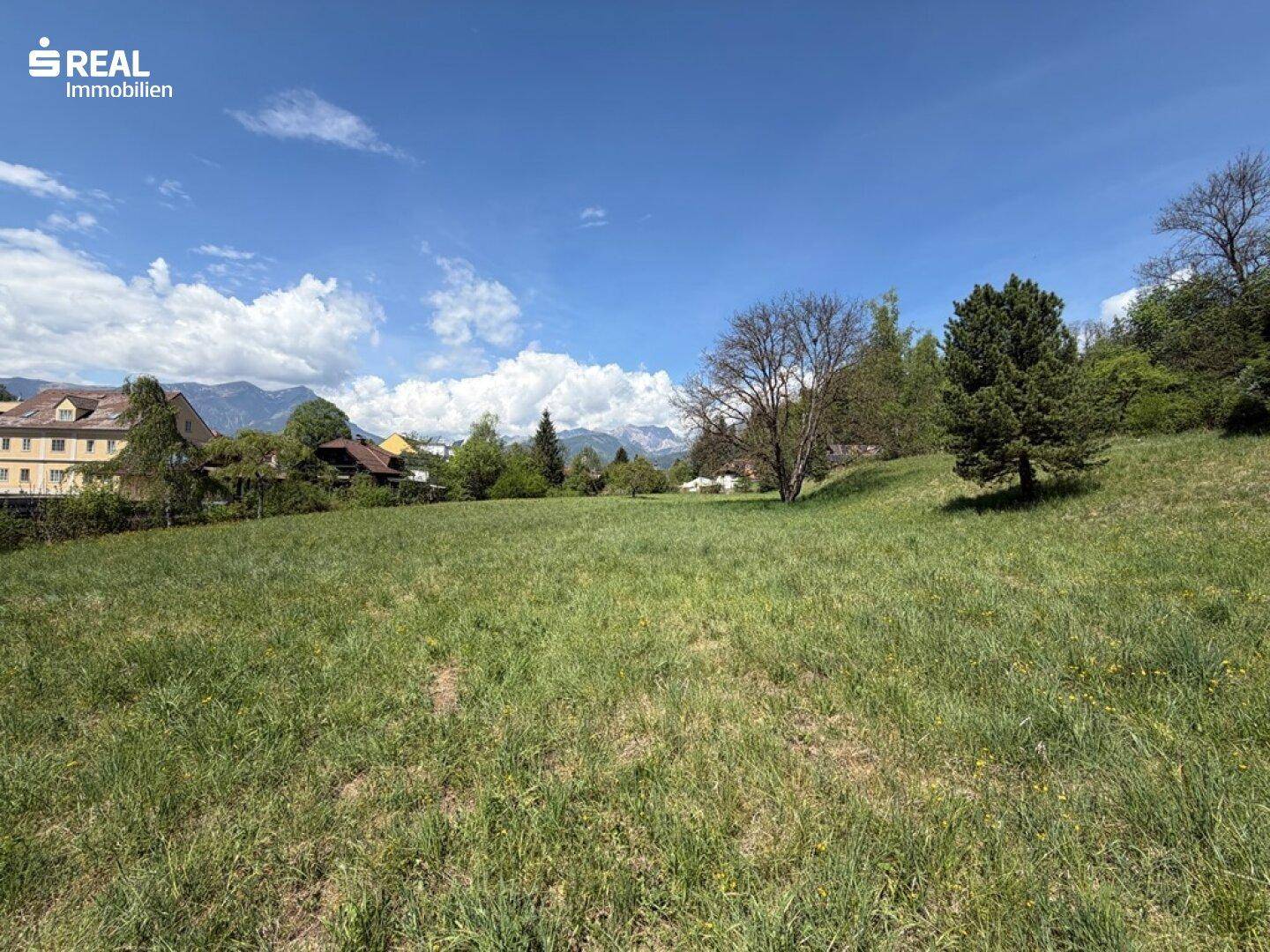 Grünes Grundstück mit Fernblick auf die Berge und umliegende Bebauung unter blauem Himmel.