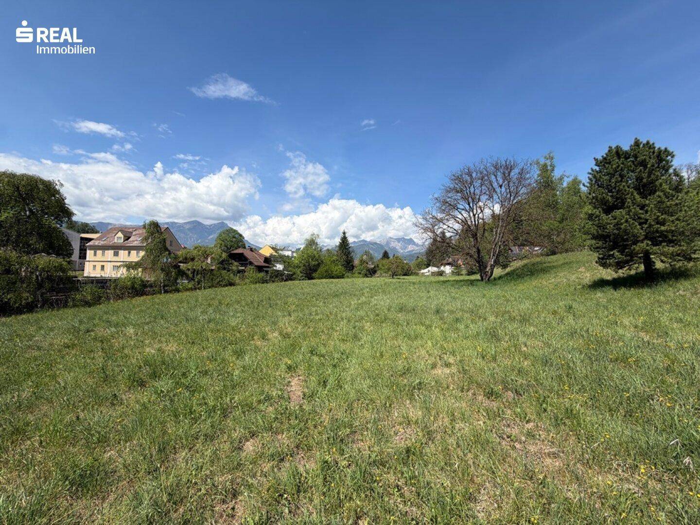 Grünes Grundstück mit Fernblick auf die Berge und umliegende Bebauung unter blauem Himmel.