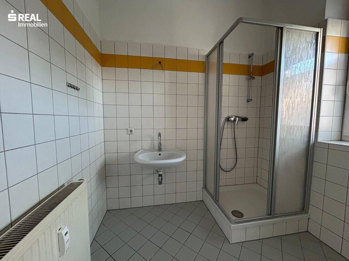 Badezimmer mit Duschkabine, Waschbecken und weiß-gelben Fliesen im Standardzustand.