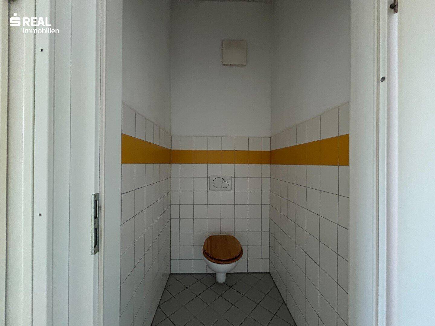 Separate Toilette mit WC, Fliesenboden und gelbem Akzentstreifen, funktional gestaltet.