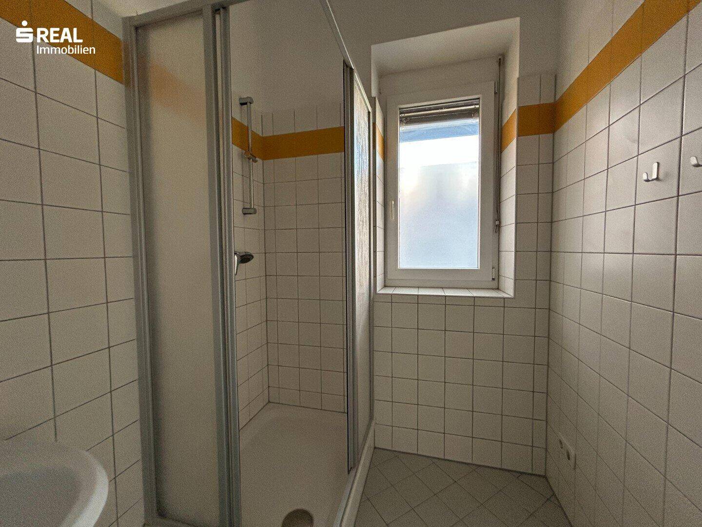 Badezimmer mit Duschkabine, Fenster und weiß-gelben Wandfliesen, gut beleuchtet.