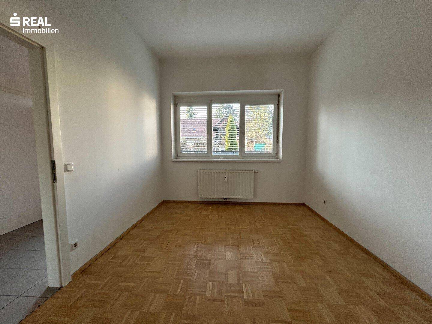Helles Schlafzimmer mit Parkettboden, großem Fenster und Heizkörper unter dem Fenster.