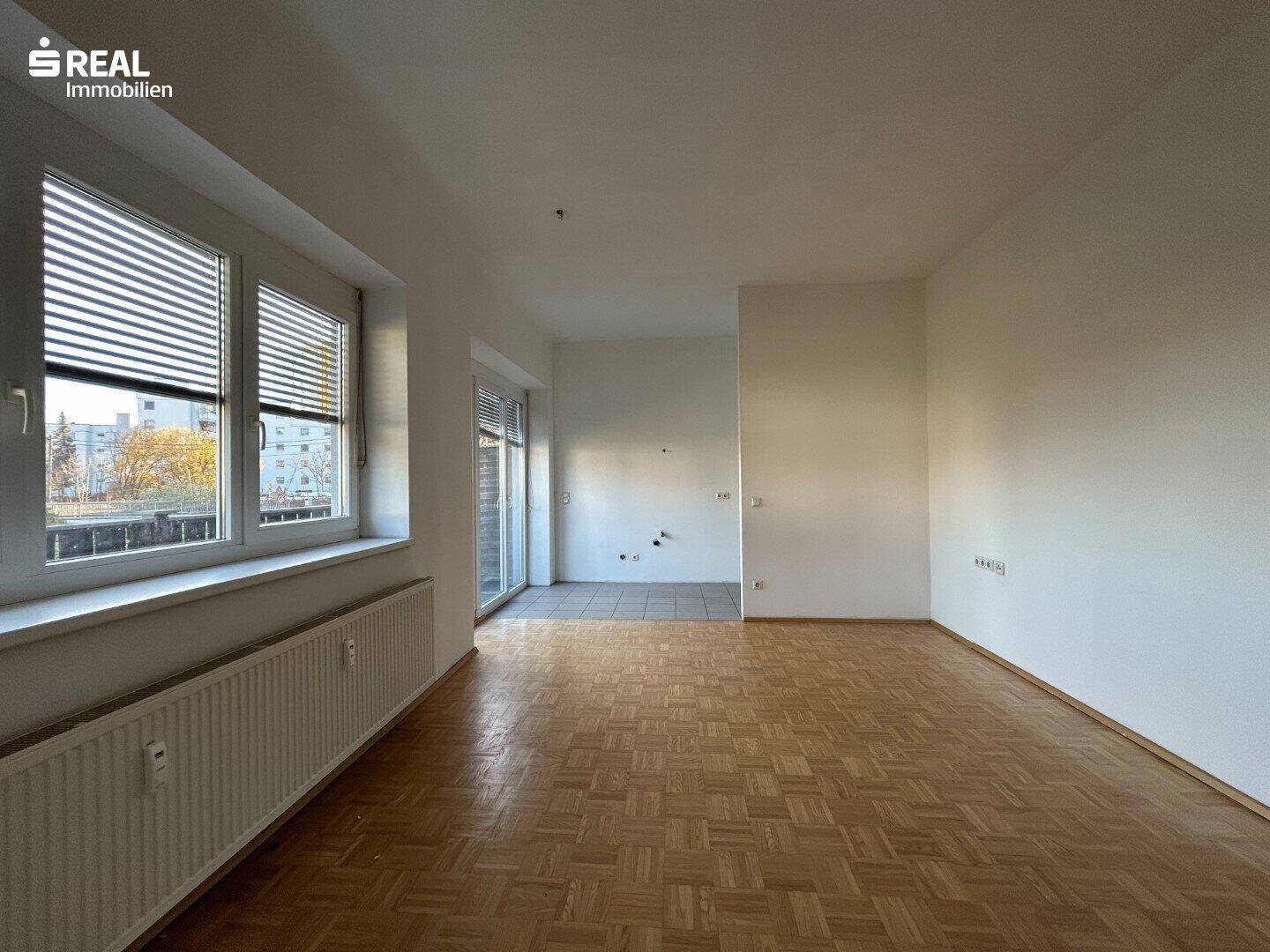 Geräumiges Wohnzimmer mit Parkettboden, großen Fenstern und Zugang zu einem weiteren Bereich.