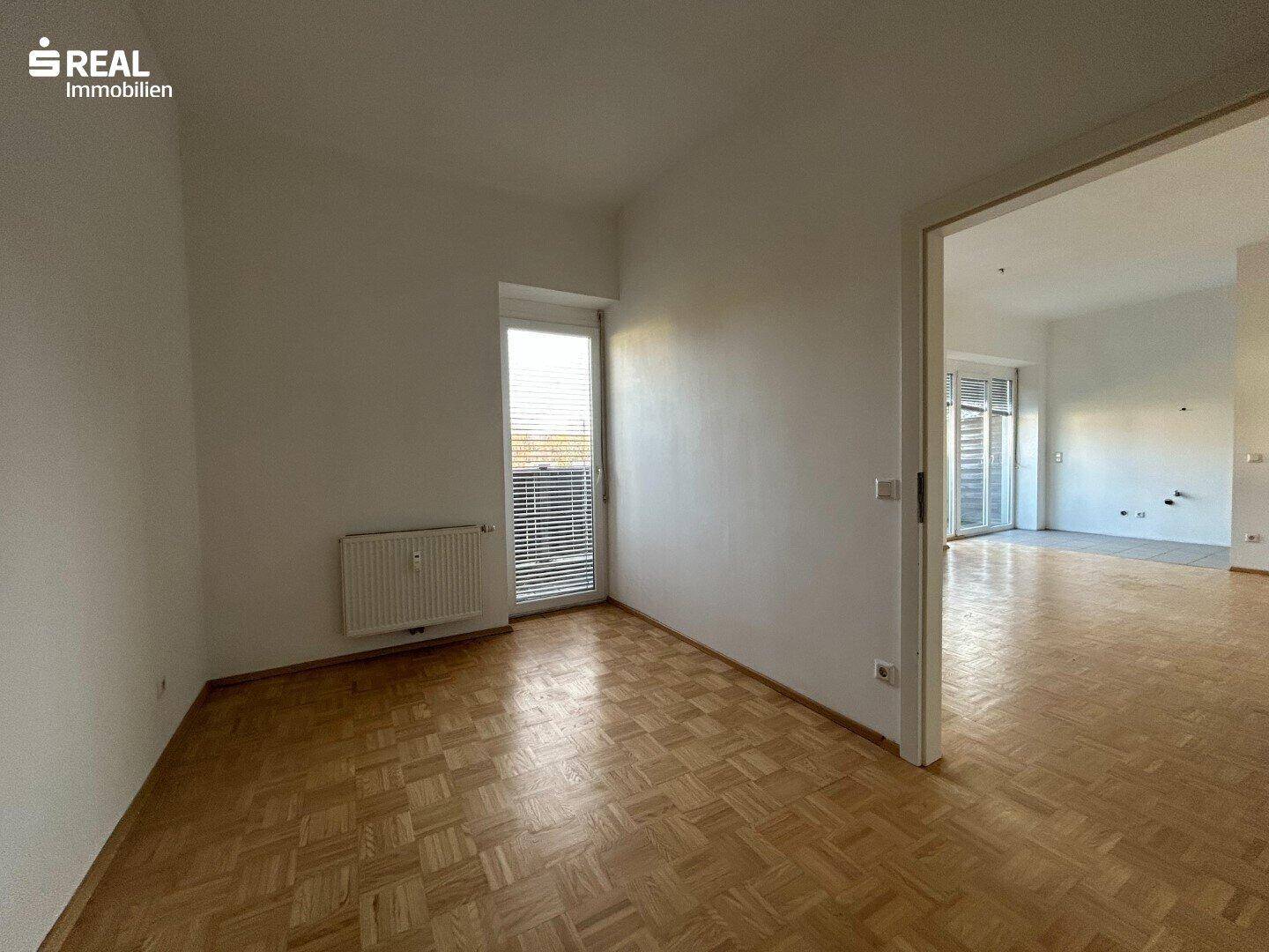 Helles Zimmer mit Parkettboden, Fenster und Zugang zu einem Balkon oder einer Terrasse.