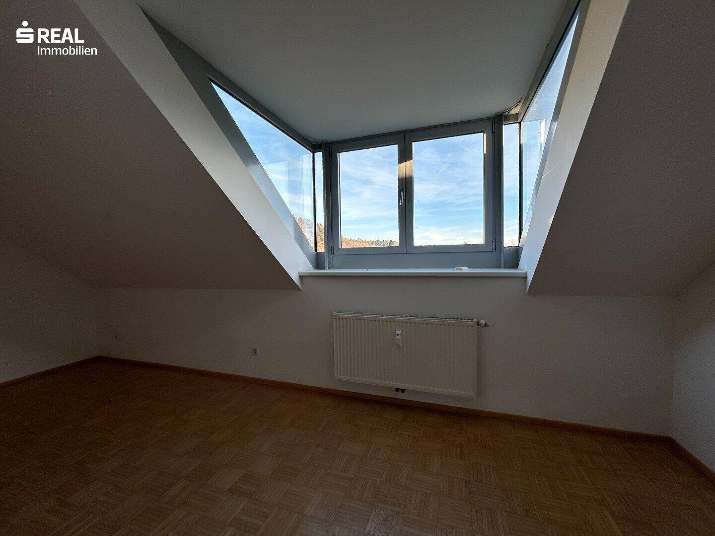 Dachraum mit Parkettboden, schrägen Wänden und einem großen Fenster mit Ausblick.