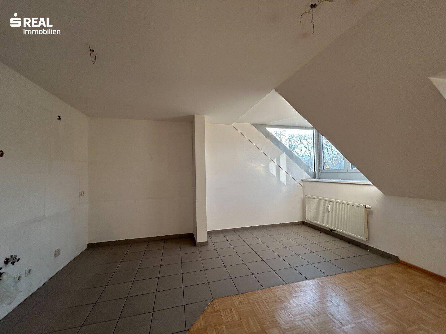 Heller Dachraum mit Fliesen- und Parkettboden, schrägen Wänden und einem großen Fenster.
