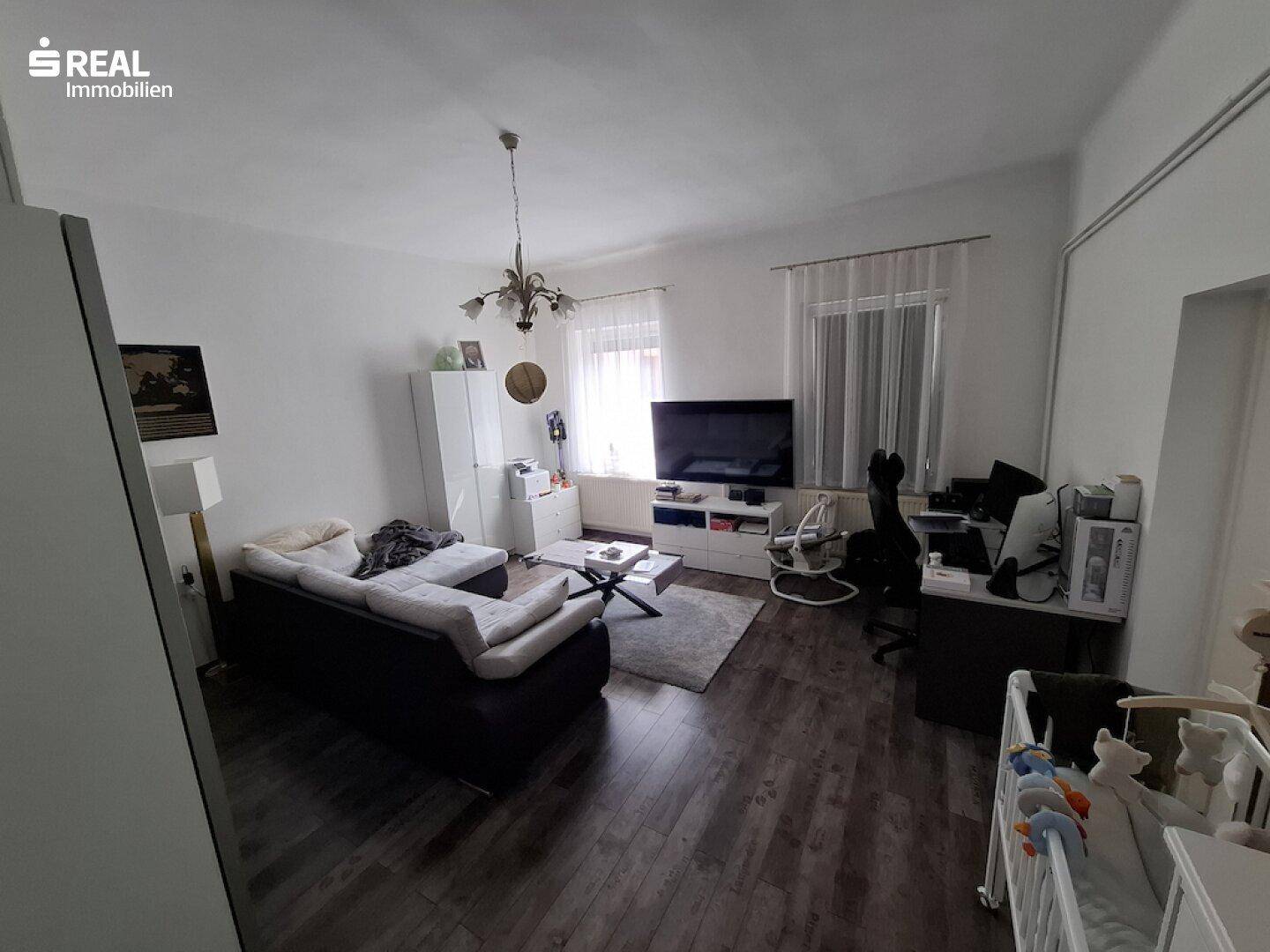 Gemütliches Wohnzimmer mit dunklem Holzboden, Sofa und einem Arbeitsbereich am Fenster.