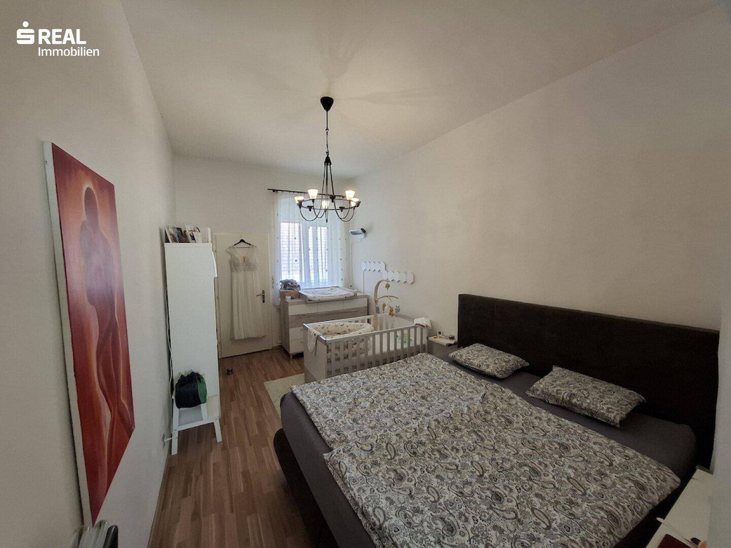 Schlafzimmer mit Doppelbett, Holzboden und einem Kinderbett am Fenster.