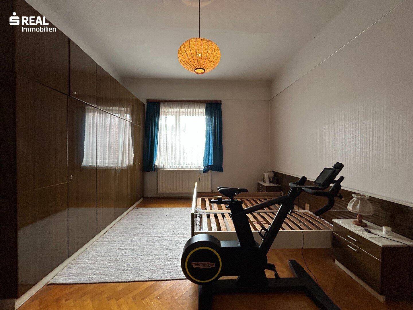 Schlafzimmer mit Holzboden, Einbauschränken und einem Trainingsgerät am Fenster.