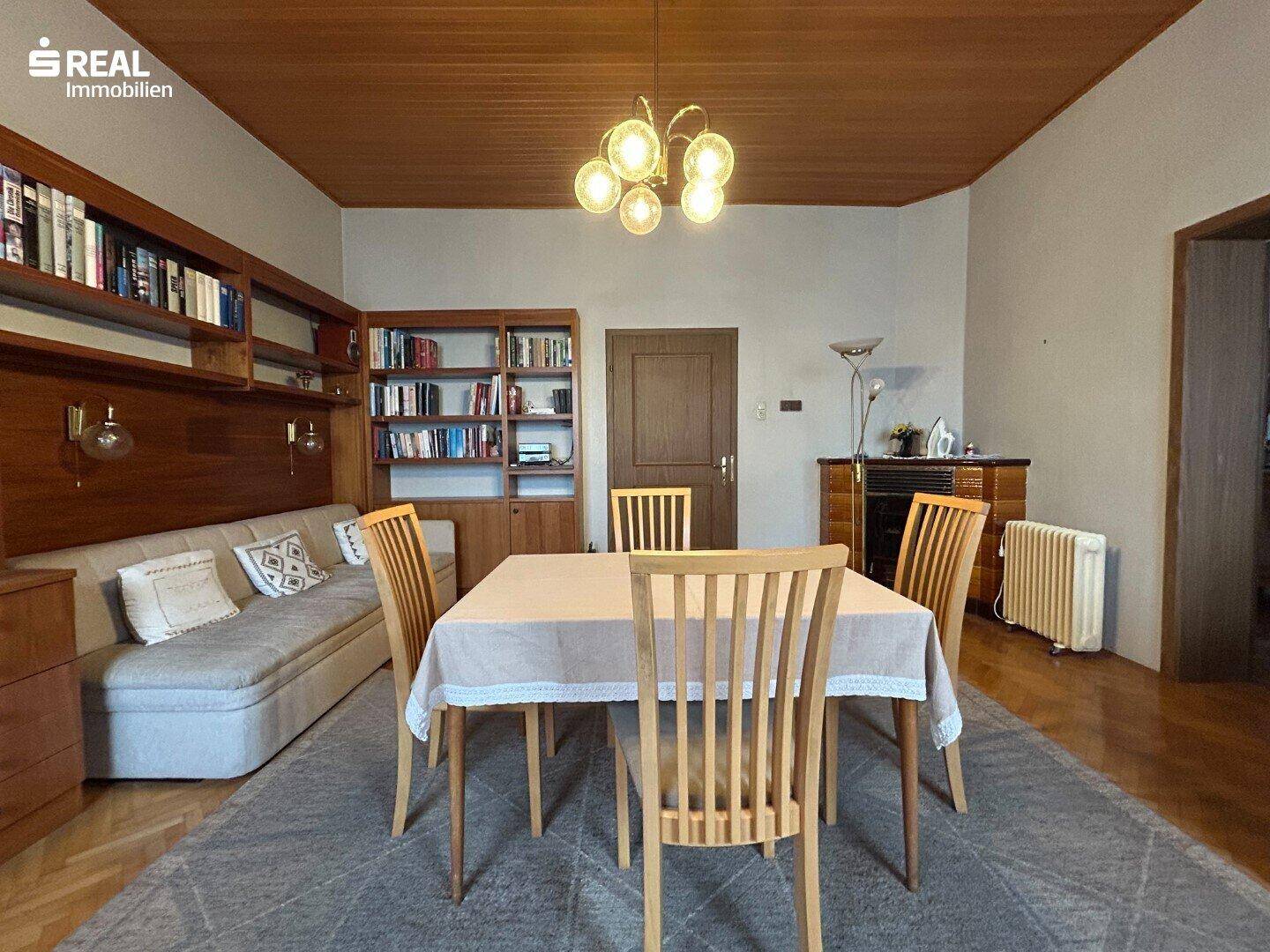 Geräumiger Wohn-Essbereich mit Holzboden, Bücherregalen und einem Kachelofen im Hintergrund.