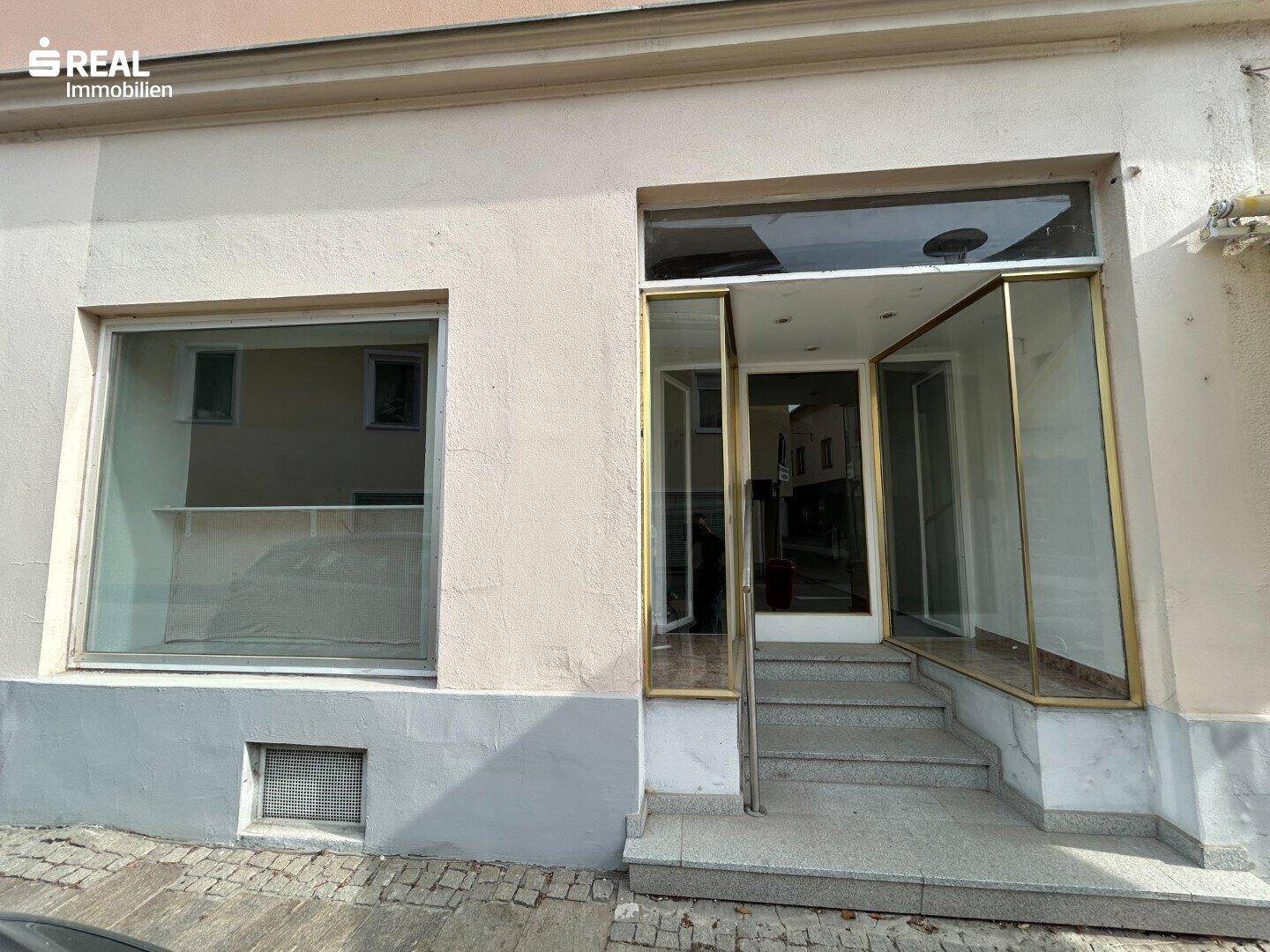 Eingangsbereich eines Geschäfts mit großen Schaufenstern und Stufen zur Straße.