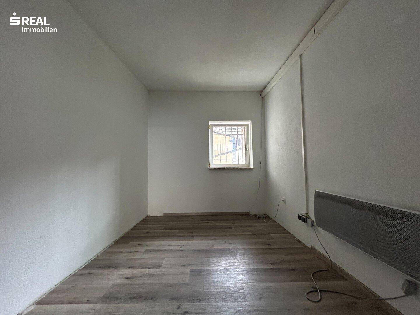 Leerer Raum mit Holzboden, weiß gestrichenen Wänden und einem Fenster mit Gitter.