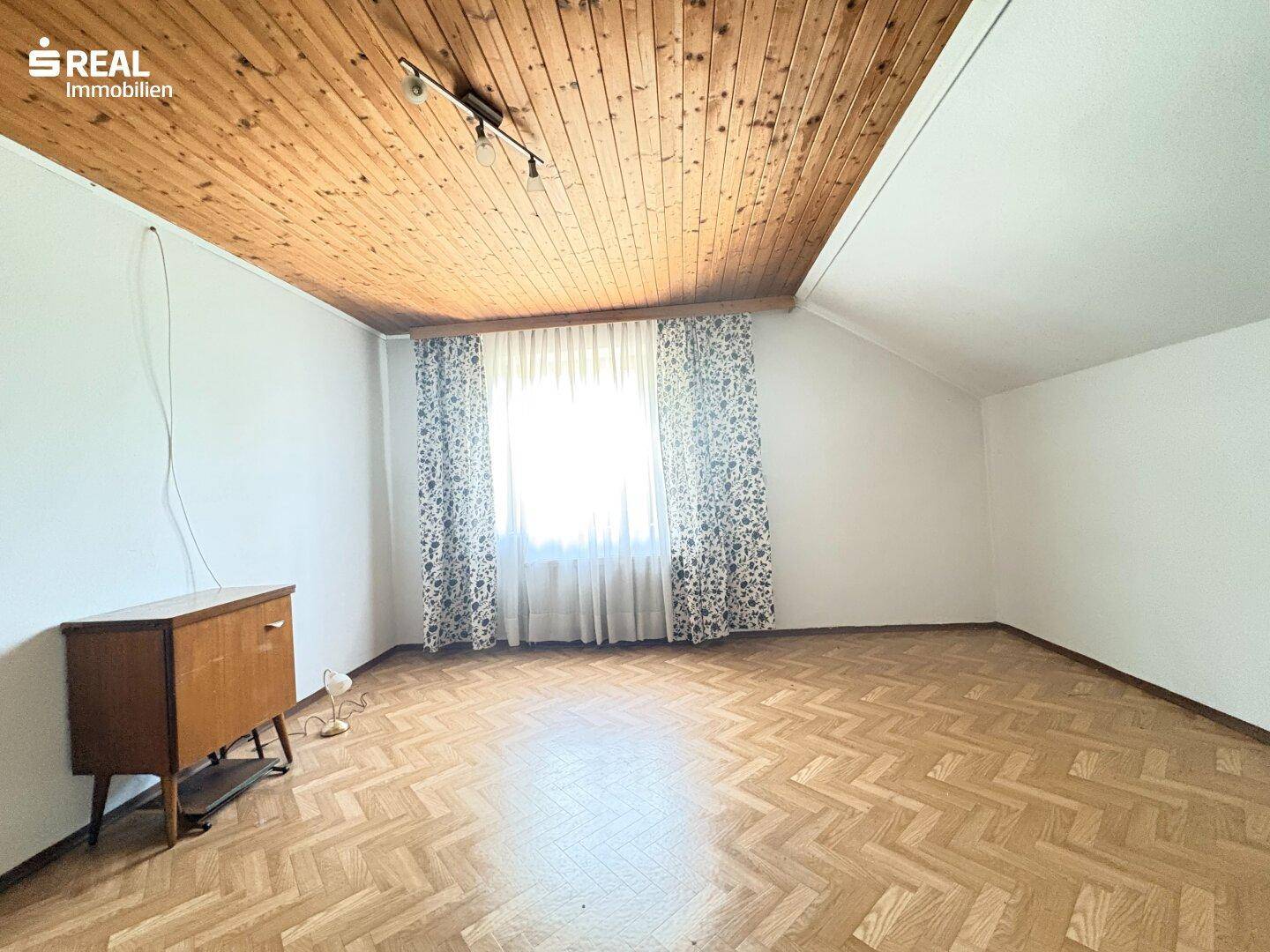 Helles Zimmer mit Fischgrätparkett, Holzdecke und einem großen Fenster mit Vorhängen.