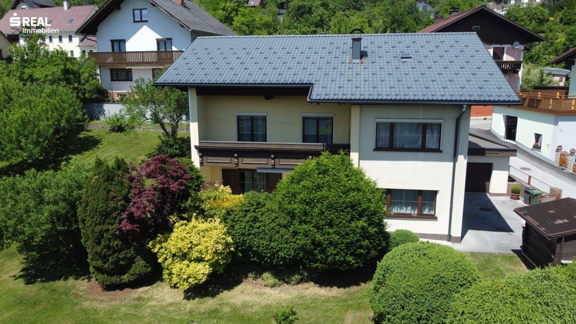 Das Haus mit seinem gepflegten Garten und der umliegenden grünen Landschaft unter blauem Himmel.