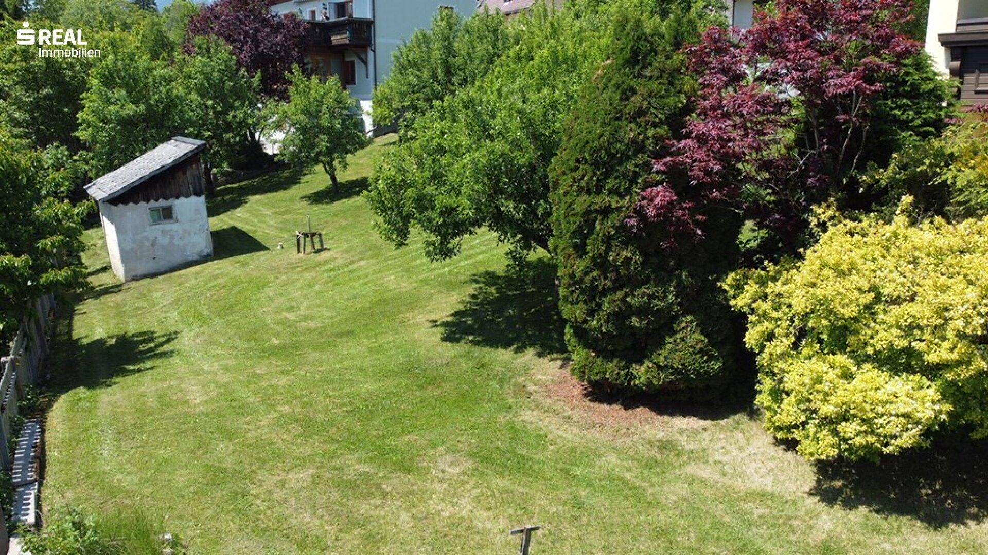 Weitläufige Rasenfläche im Garten mit einem kleinen Gartenhaus und altem Baumbestand.