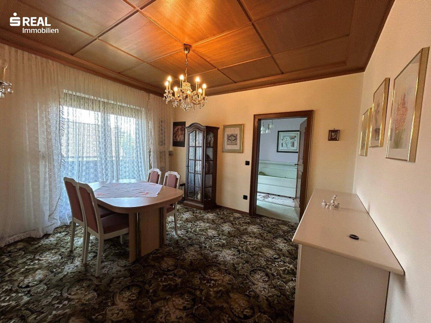 Klassisches Esszimmer mit rundem Esstisch, Vitrine und gemustertem Teppichboden unter Holzbalkendecke.