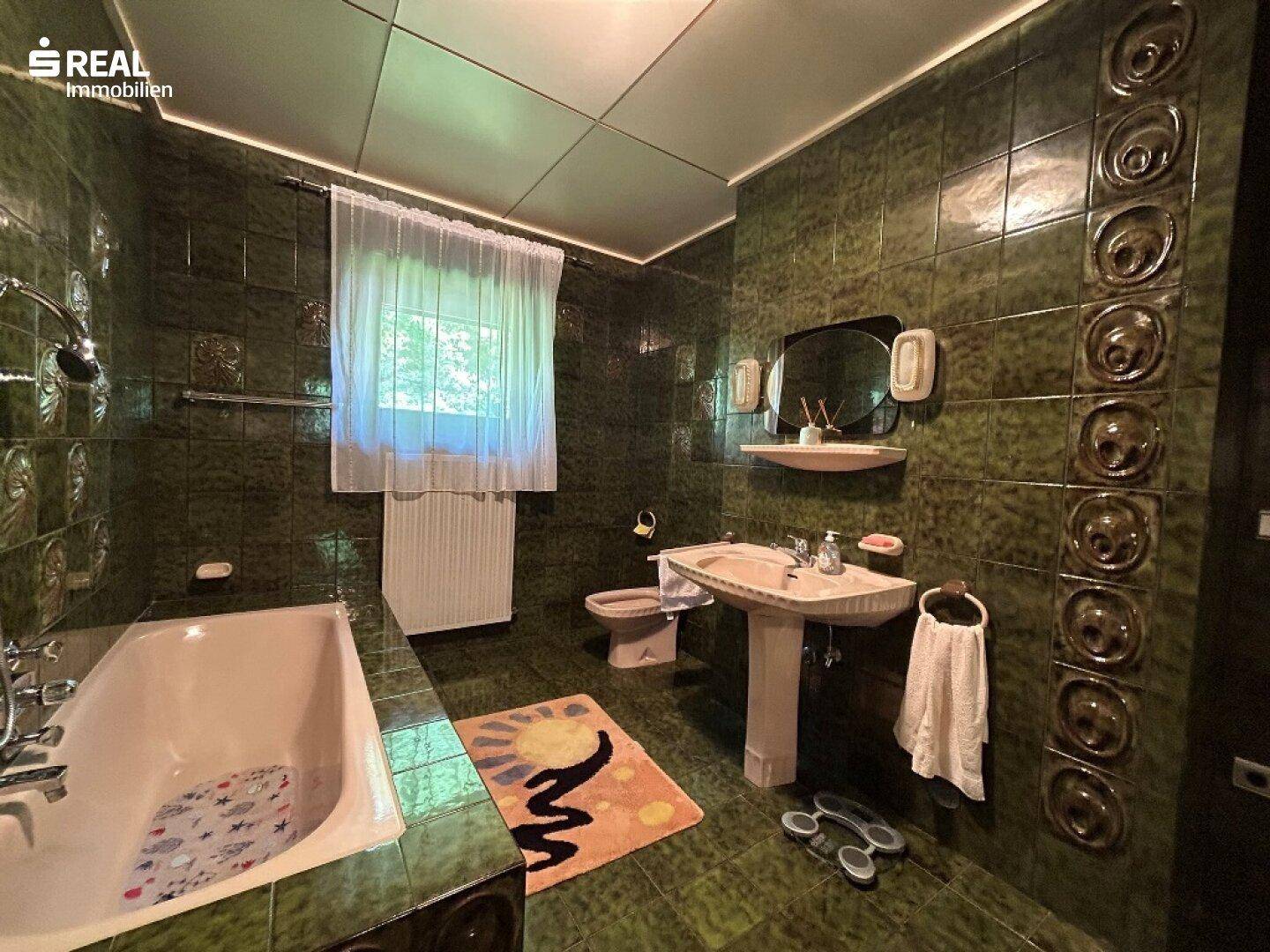 Badezimmer mit dunkelgrünen Fliesen, Badewanne, Standwaschbecken und Fenster für natürliche Belüftung.