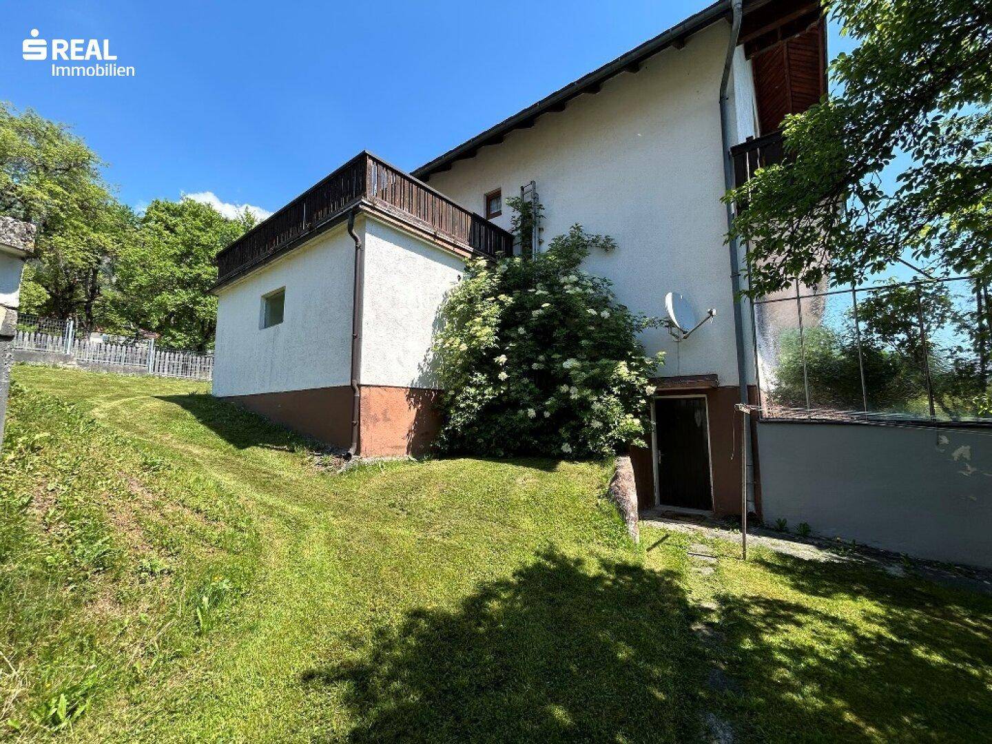 Seitenansicht des Hauses mit Zugang zum Keller und einem Hanggrundstück, umgeben von üppigem Grün.