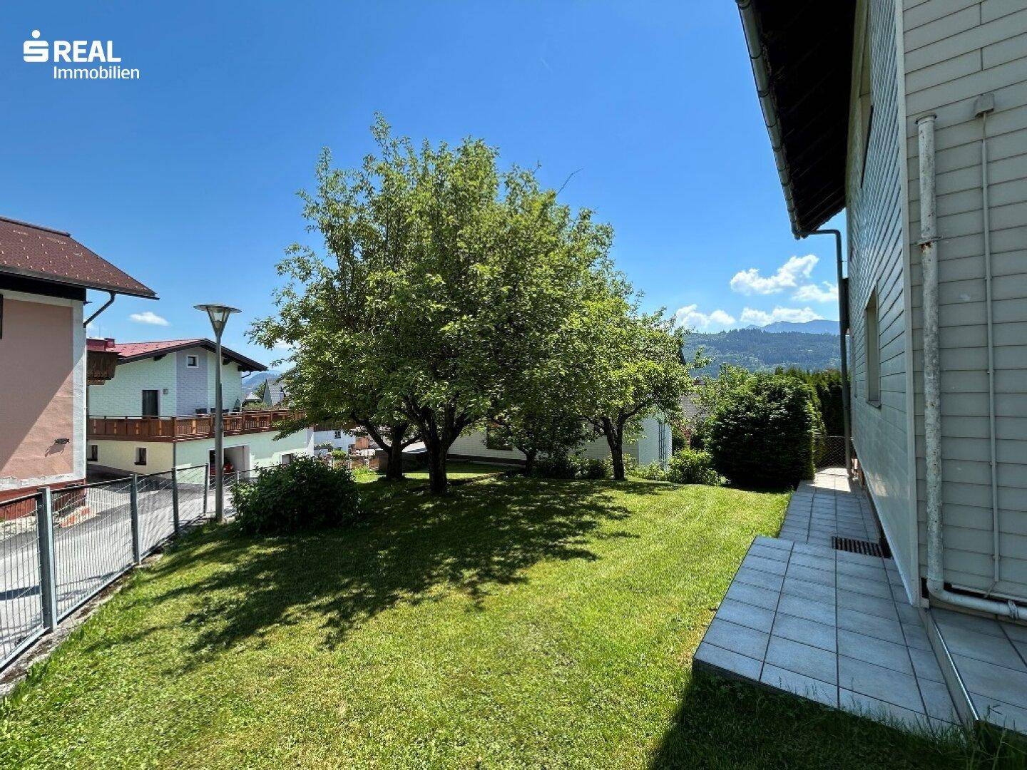 Gepflegter Garten mit Bäumen und einer gefliesten Fläche neben dem Haus, ideal für Outdoor-Aktivitäten.