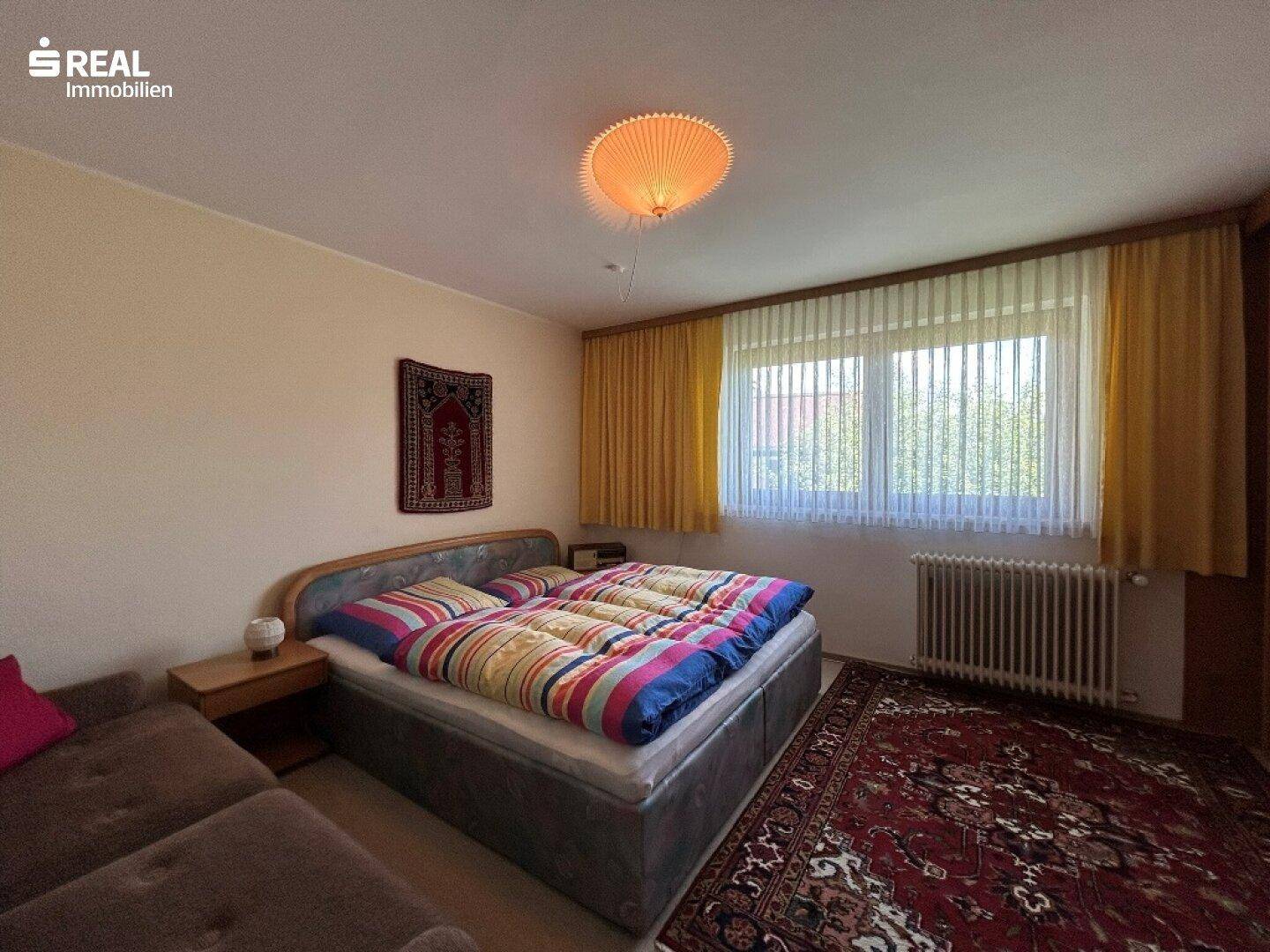Gemütliches Schlafzimmer mit Doppelbett, Teppichboden und gelben Vorhängen, mit Blick ins Grüne.