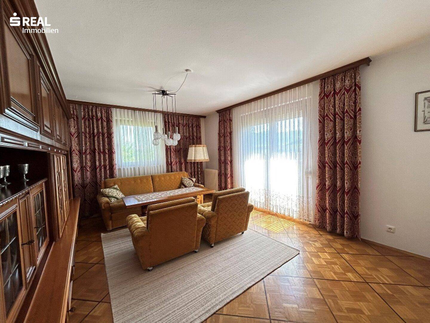Traditionelles Wohnzimmer mit Parkettboden, Holzmöbeln und großen Fenstern mit Vorhängen.