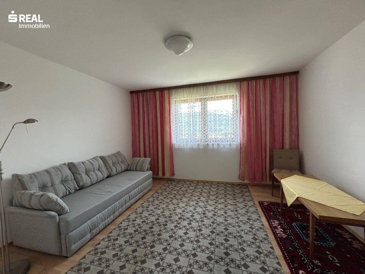 Helles Zimmer mit Sofa, Teppich und Fenster, das als Wohn- oder Schlafzimmer genutzt werden kann.