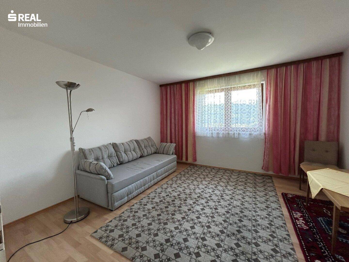 Einfaches Wohnzimmer mit hellem Laminatboden, grauem Sofa und rosa Vorhängen.