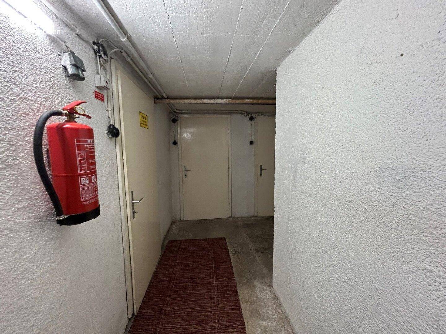 Kellerflur mit Betonwänden, Feuerlöscher und Zugang zu mehreren Kellerräumen.