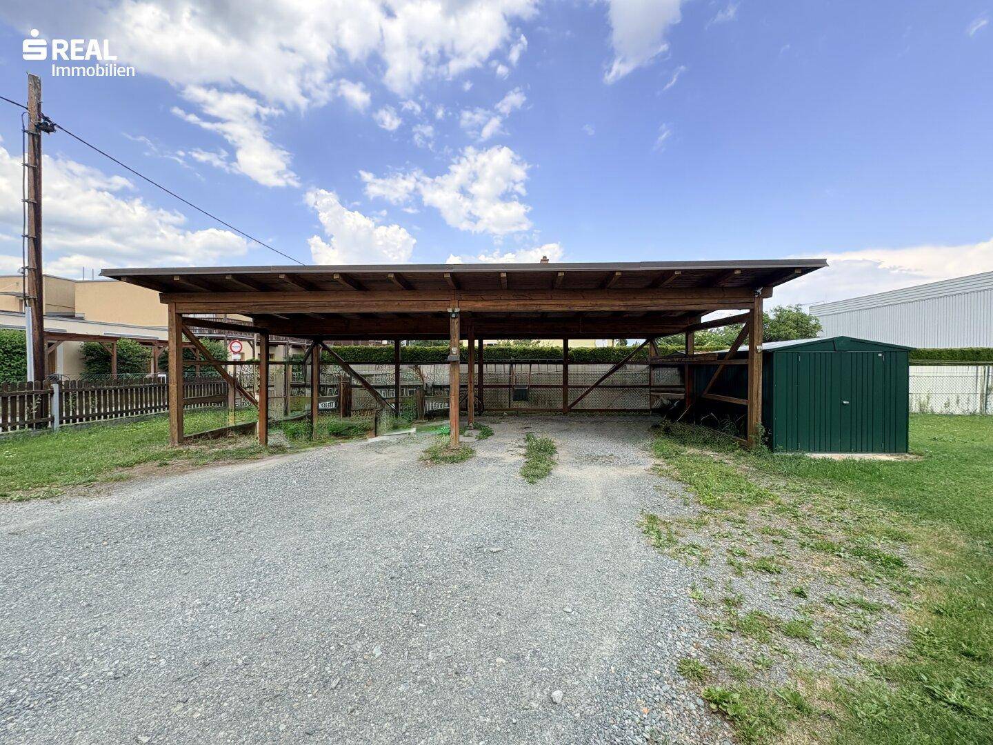 Geräumiger Holzcarport mit Schotterfläche und einem grünen Gartenschuppen auf dem Grundstück.