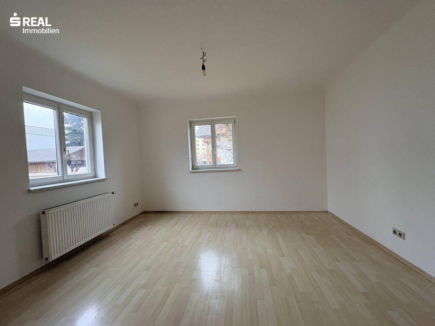 Leeres Zimmer mit Laminatboden, zwei Fenstern und einem Heizkörper unter einem Fenster.