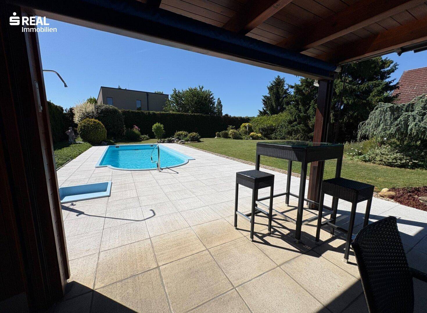 Überdachte Terrasse mit Sitzgelegenheiten und direktem Blick auf den Pool und den Garten.