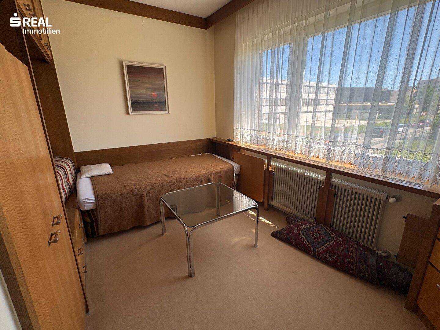 Helles Schlafzimmer mit Einzelbett, Teppichboden, großem Fenster und einfacher Holzmöblierung.
