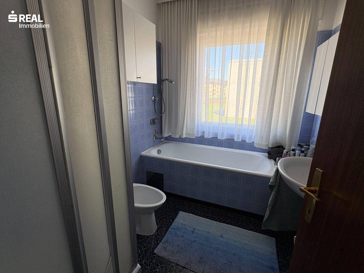 Badezimmer mit blauen und weißen Fliesen, Badewanne mit Duschfunktion, WC und Fenster.