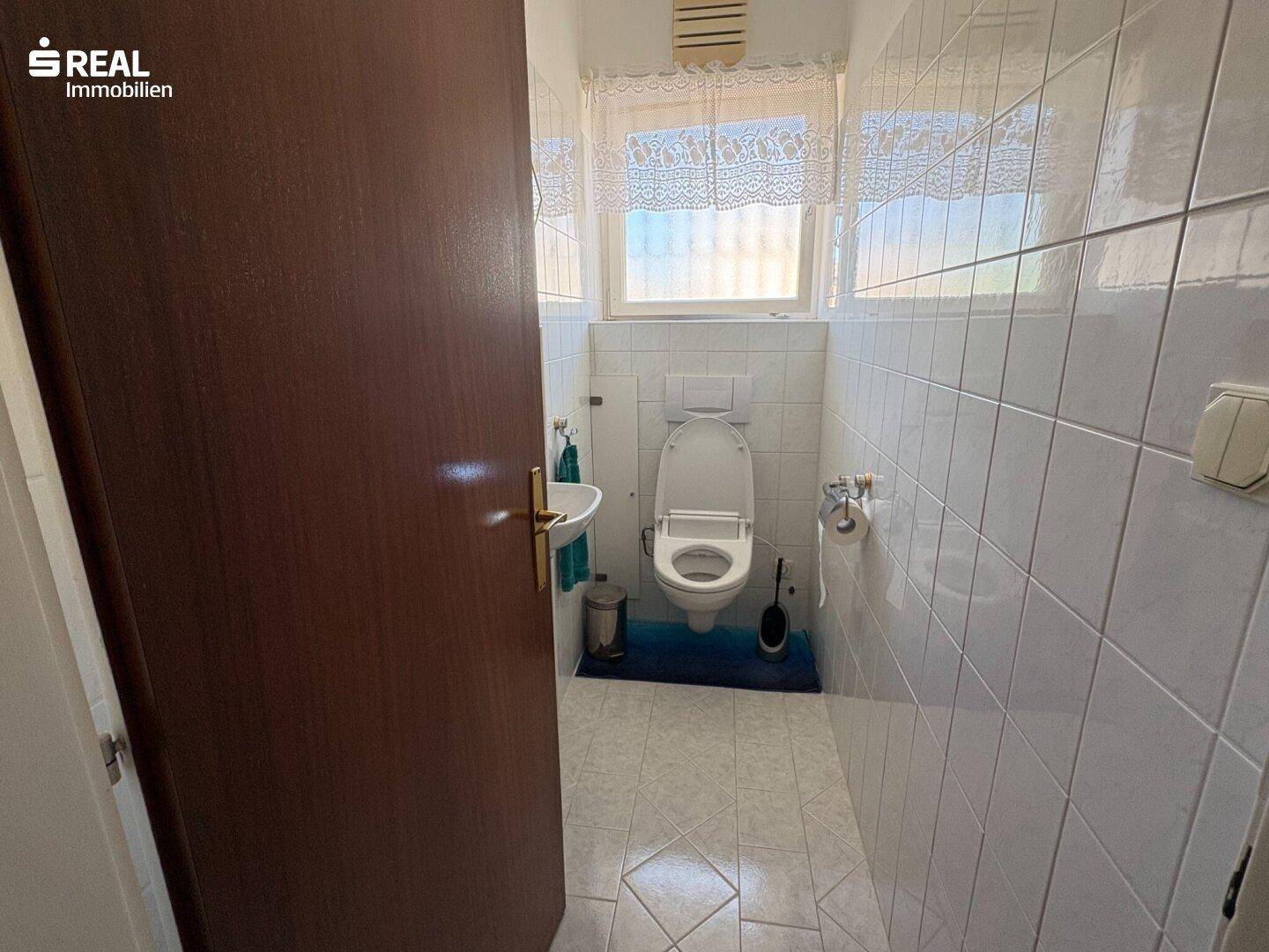Gäste-WC mit weißen Fliesen, Toilette, kleinem Waschbecken und einem Fenster für Tageslicht.
