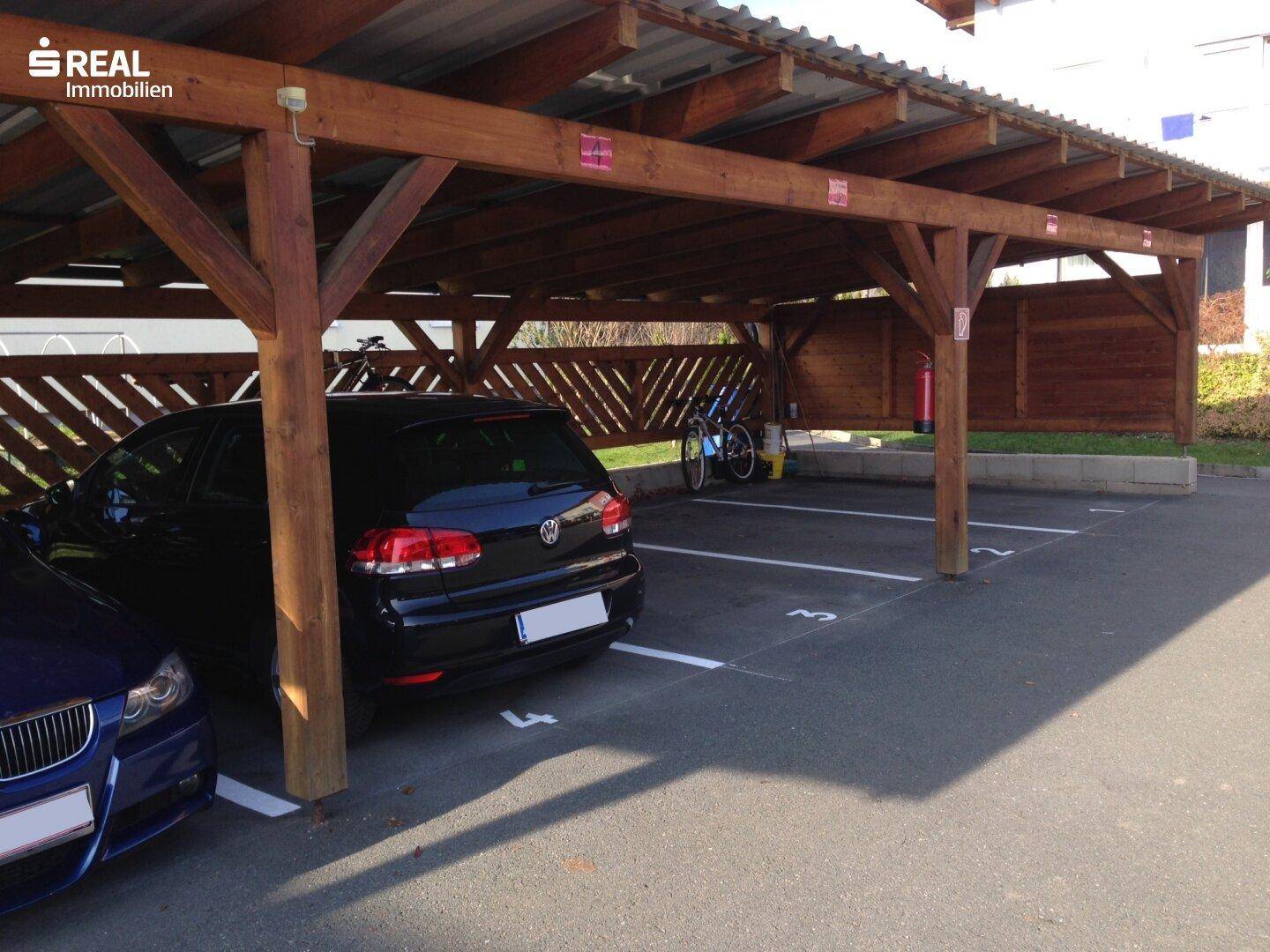 Überdachter Holzcarport mit markierten Stellplätzen und geparkten Fahrzeugen.