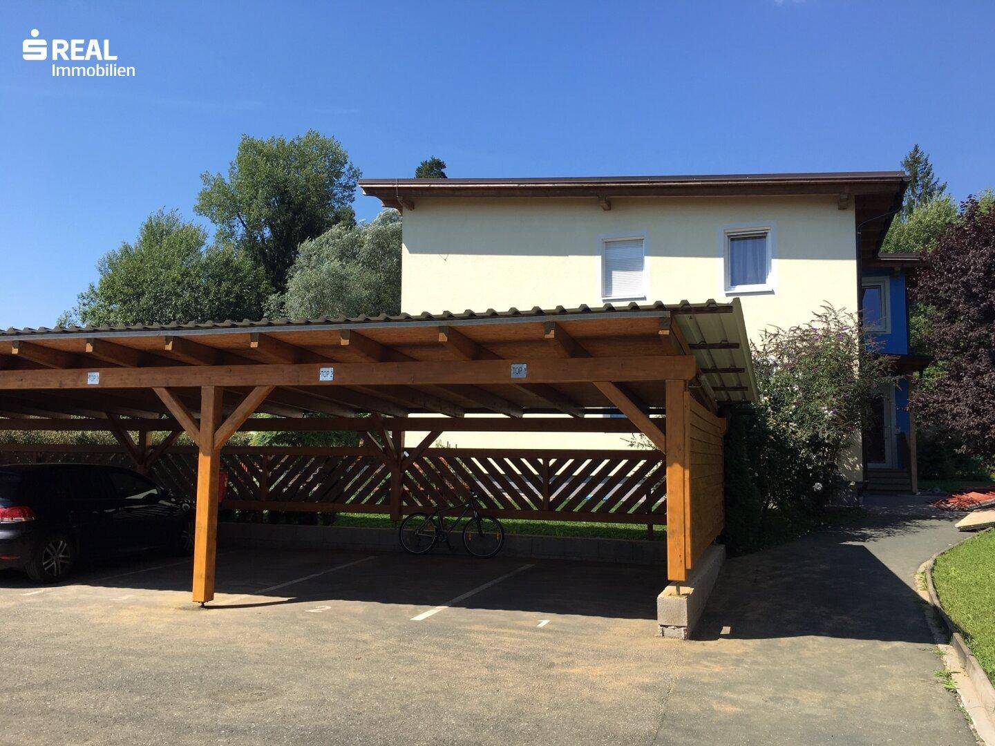 Holzcarport mit mehreren Stellplätzen vor dem Gebäude, umgeben von Grünflächen und Bäumen.