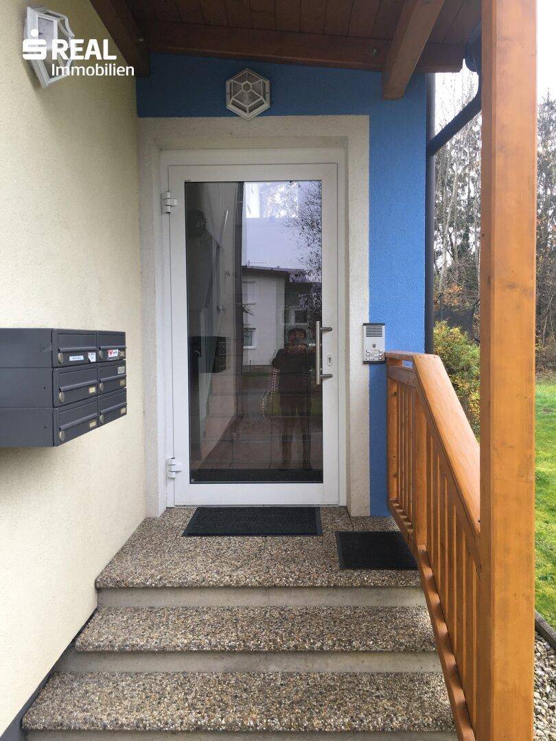 Eingangsbereich des Gebäudes mit moderner Glastür, Briefkästen und Holzgeländer.