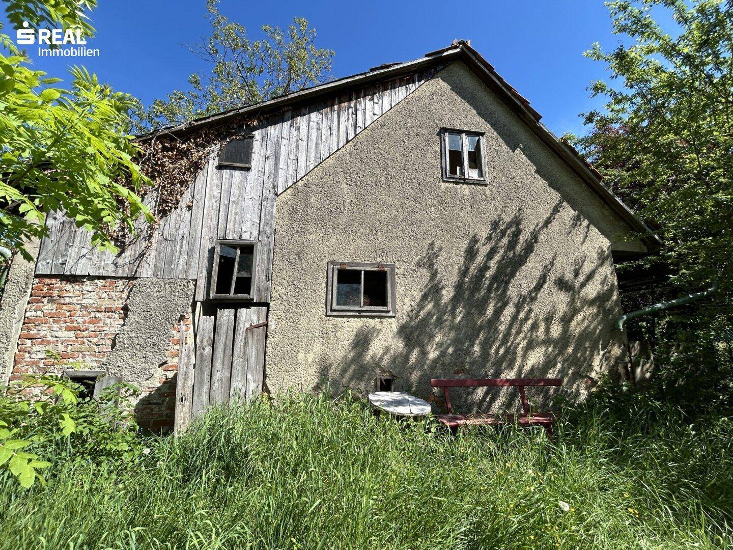 Rustikales Nebengebäude mit verwitterter Fassade, umgeben von hohem Gras und wilder Vegetation.