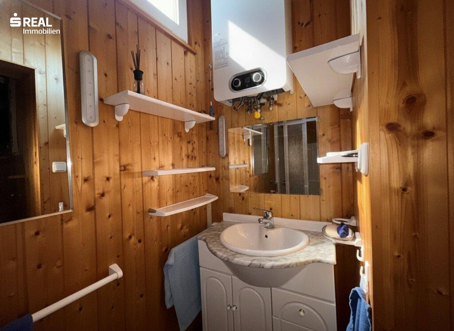 Badezimmer mit Holzverkleidung, Waschtisch, Spiegel und einem Warmwasserbereiter an der Wand.