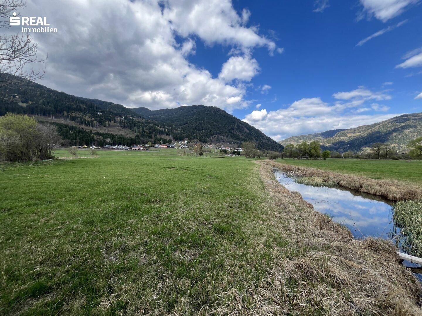 Bachufer mit grünen Wiesen und einem Dorf in der Ferne, umgeben von bewaldeten Bergen.