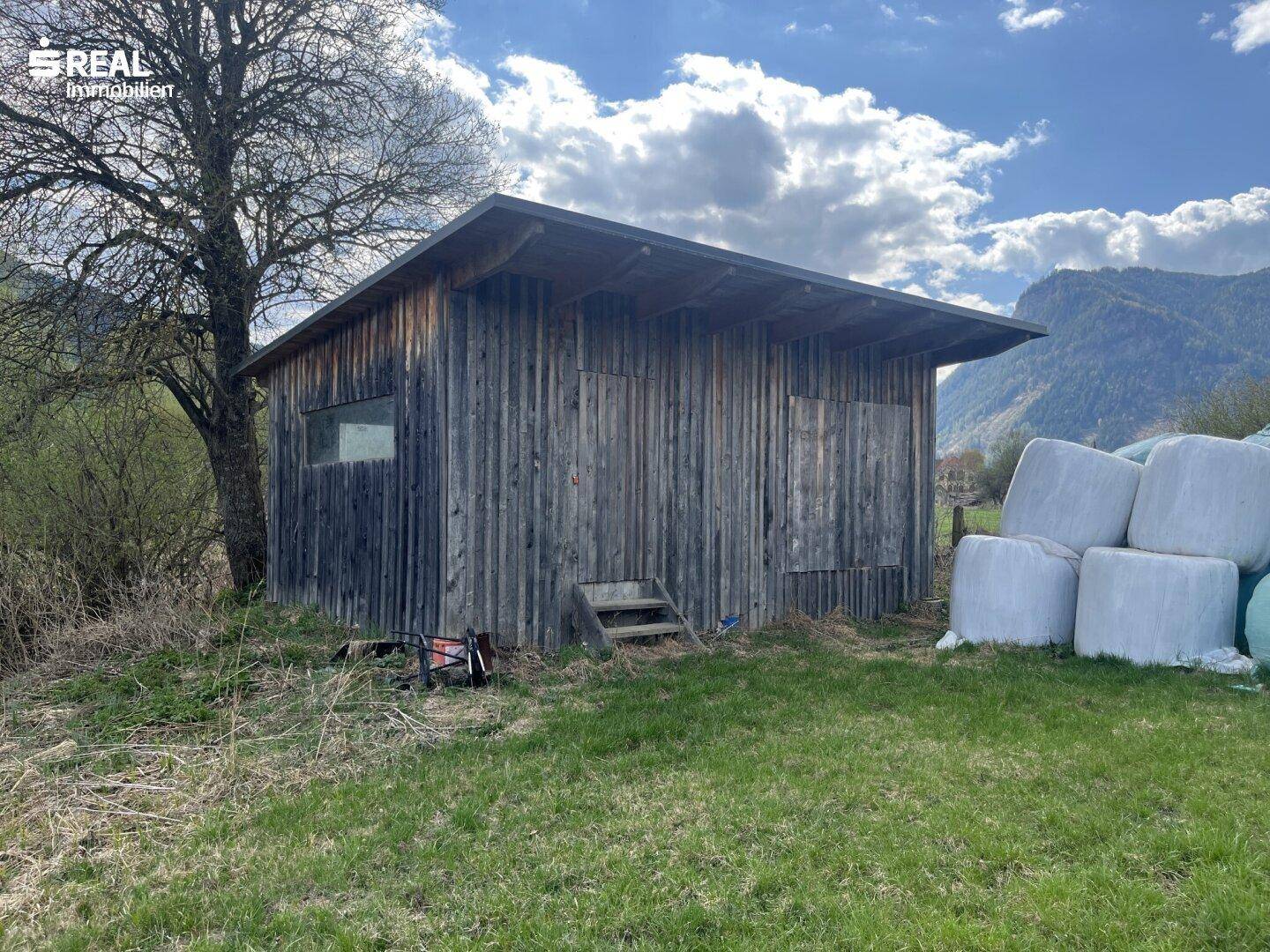 Rustikale Holzhütte neben Heuballen auf einer Wiese mit Bergen im Hintergrund.