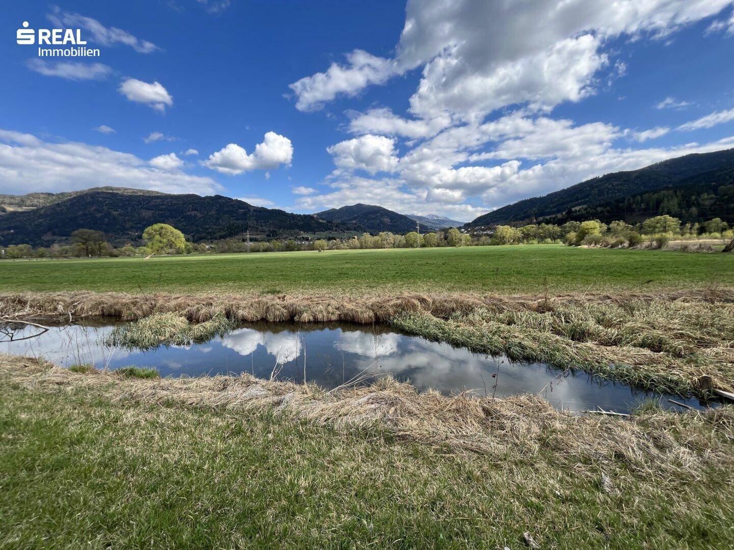 Bachlauf mit Spiegelungen des Himmels, umgeben von grünen Wiesen und bewaldeten Bergen.