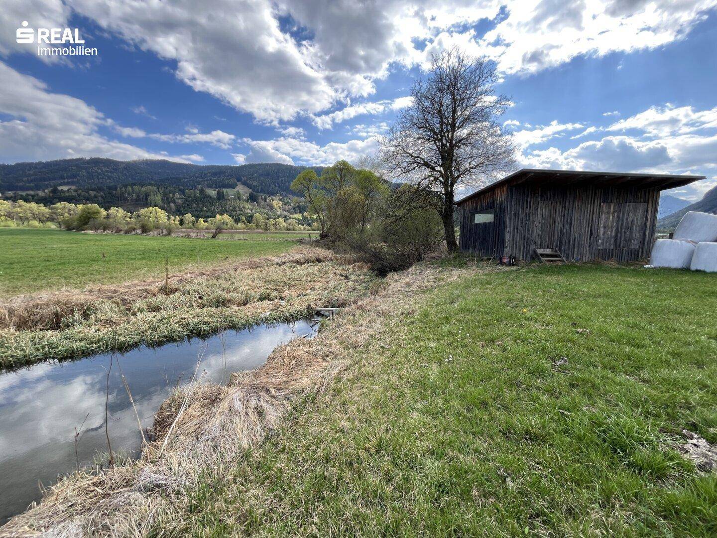 Kleiner Bachlauf neben einer Holzhütte und Heuballen, umgeben von grünen Wiesen und Bergen.