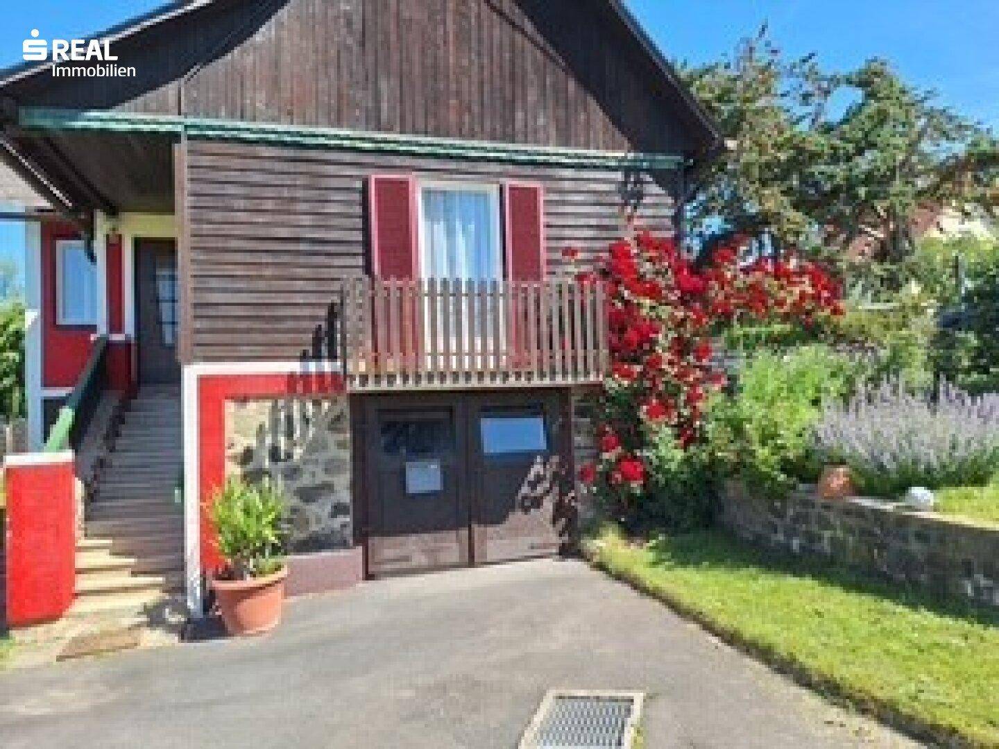 Rustikales Holzhaus mit Balkon, Garage und blühendem Gartenbereich unter blauem Himmel.