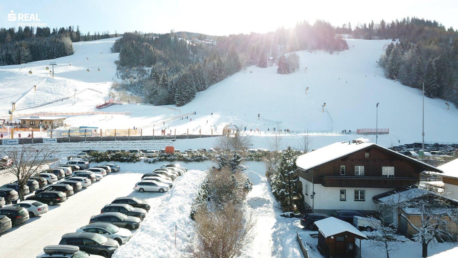 Winterliche Szenerie mit Haus, Parkplatz und belebter Skipiste im Hintergrund.