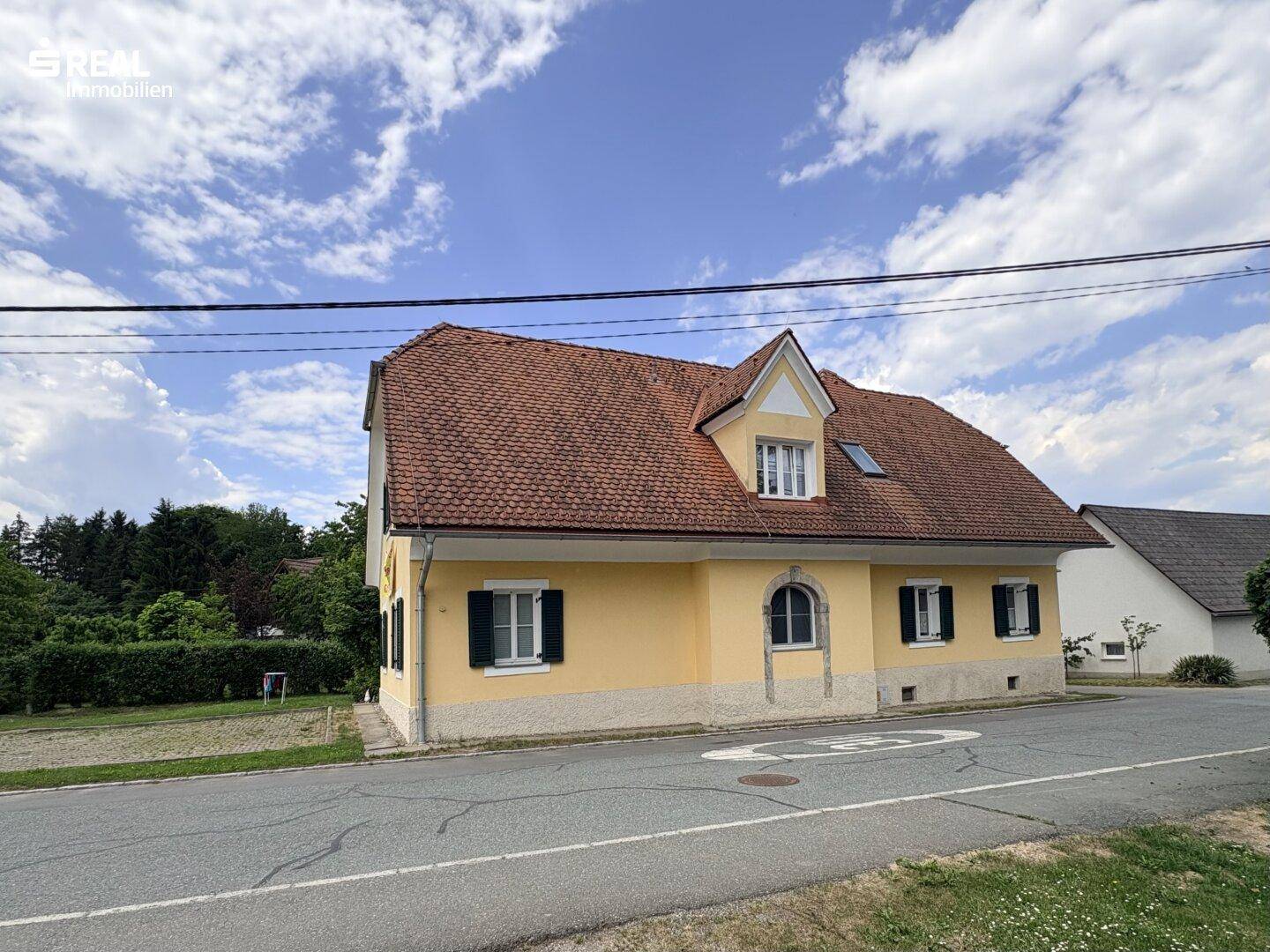 Ansicht des gelben Hauses von der Straßenseite mit rotem Ziegeldach und grünen Fensterläden.