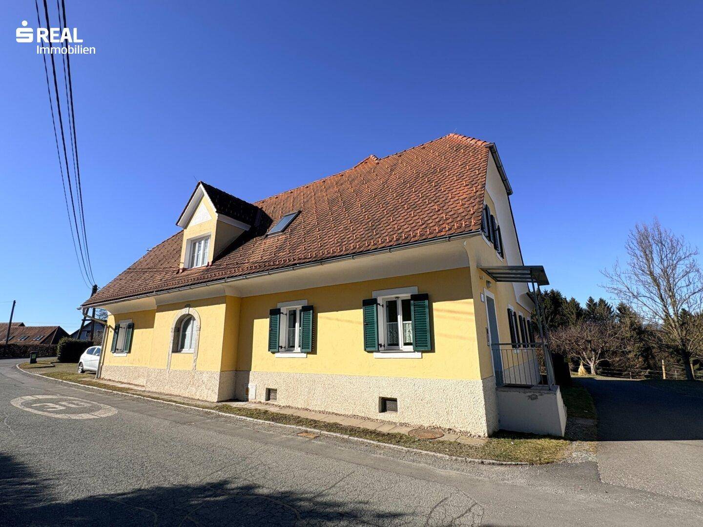 Seitenansicht des gelben Hauses mit rotem Ziegeldach, grünen Fensterläden und kleinem Balkon.