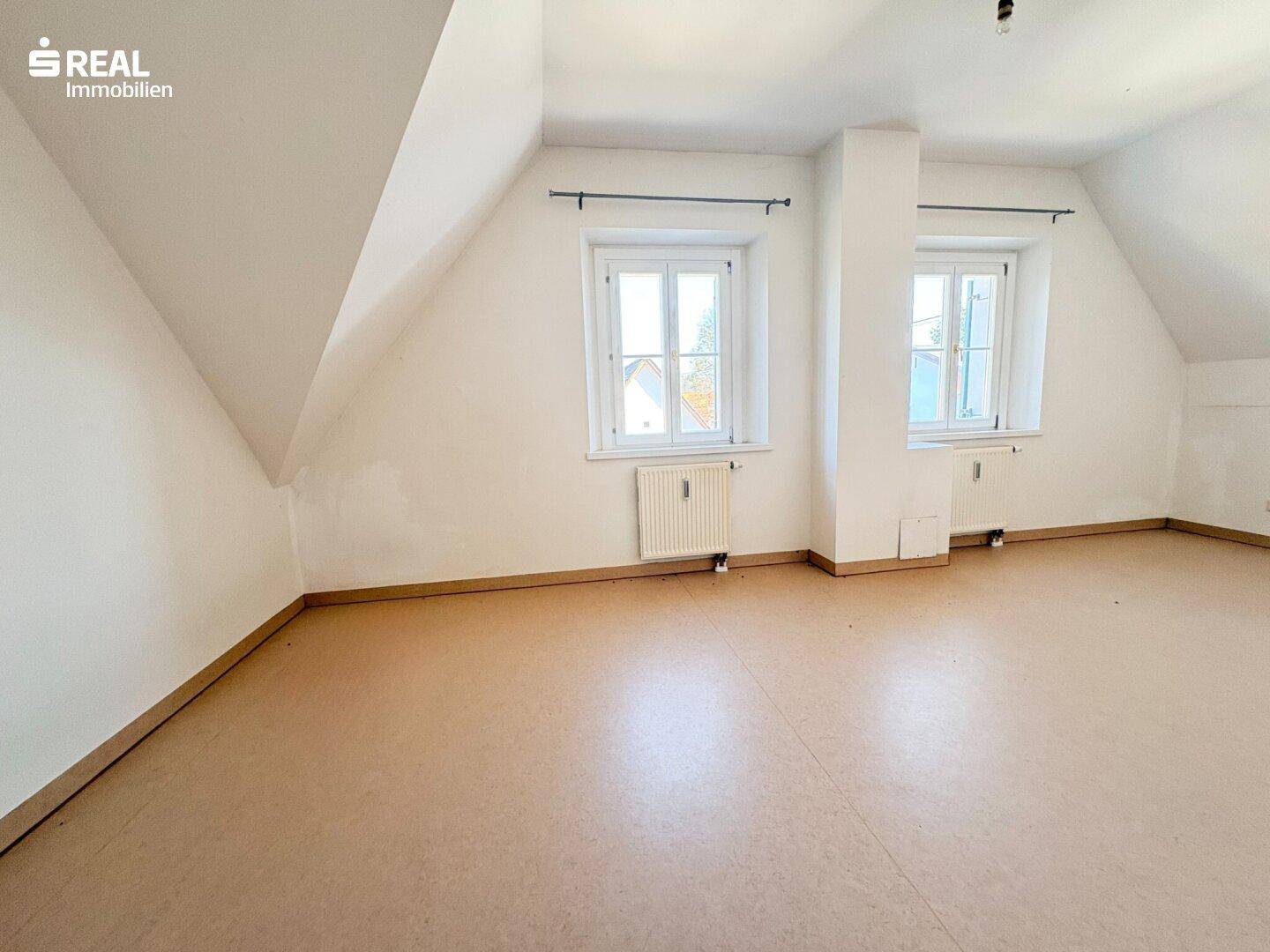 Geräumiges Zimmer im Dachgeschoss mit zwei Fenstern, Heizkörpern und Dachschrägen.