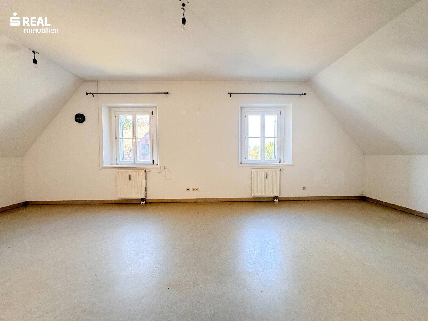Helles Dachgeschosszimmer mit zwei Fenstern, weiß gestrichenen Wänden und Linoleumboden.