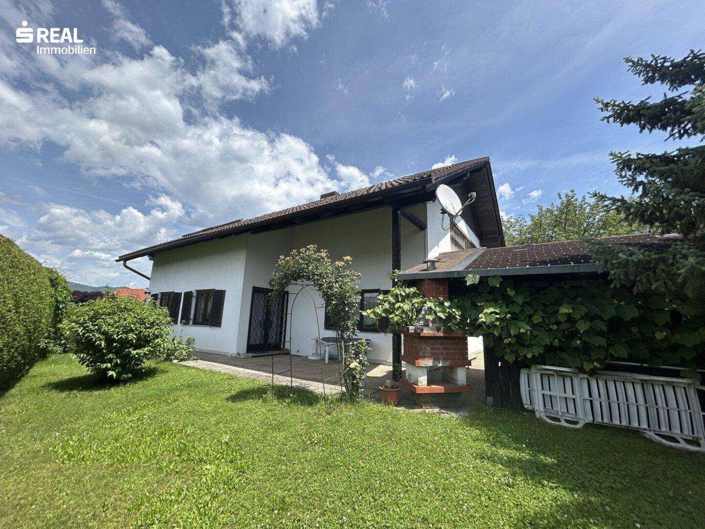 Außenansicht des Hauses mit gepflegtem Garten und Grillplatz im Freien unter blauem Himmel.